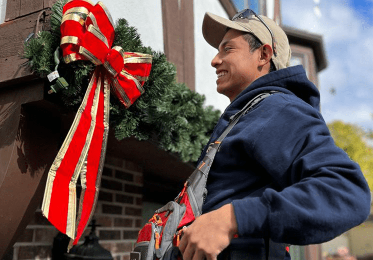 A worker smiling while installing a large holiday wreath with red ribbons on a home exterior.