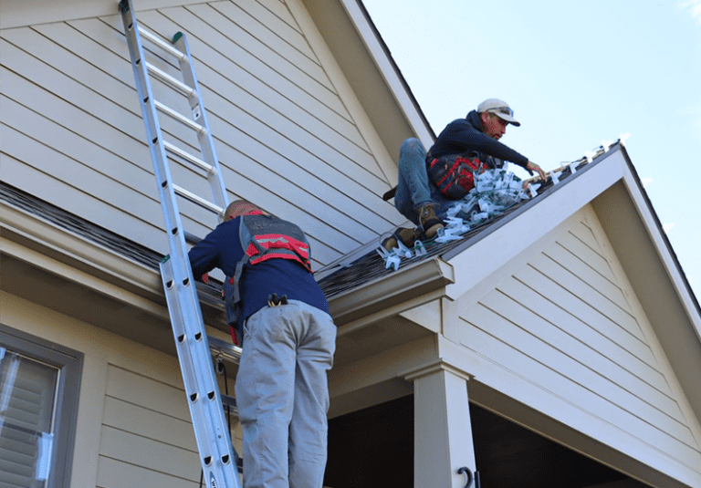 Two workers on a ladder and roof installing holiday lights on a home